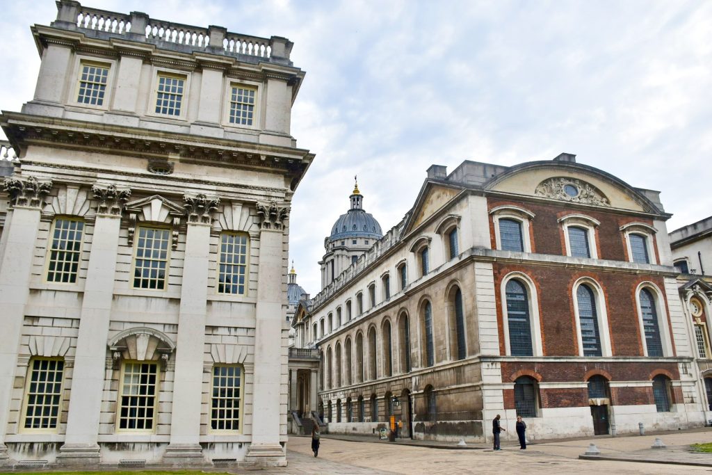 Old fashioned building with walkway in-between. To the left a white stone building with square roof and rows of three windows over three flours. To the right is a white stone building with a red brick end. It looks potentially like a church given the arched windows around the building.