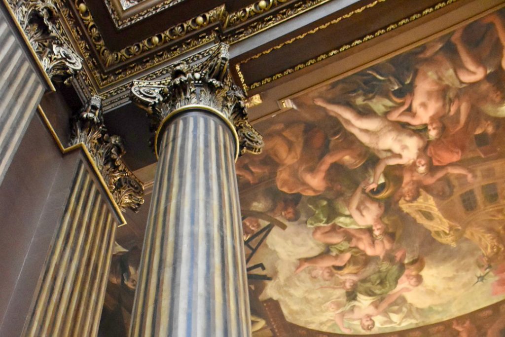 Internal photo looking up at a ceiling painted with an old fashioned painting. There is a striped colour to the left in browns and golds