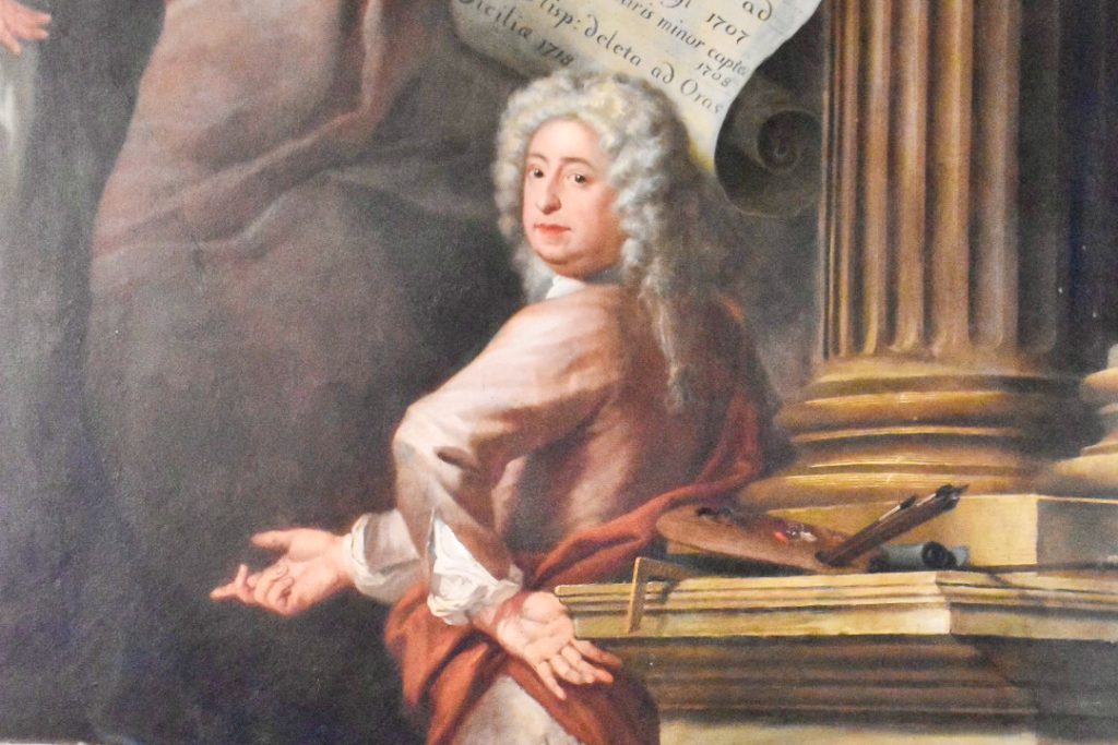 Painting of a man in period dress with white wig and pink frock coat in front of a gold pillar. A painting easel behind him. His left hand is on his hip and his right hand pointing forwards. He is looking at the camera