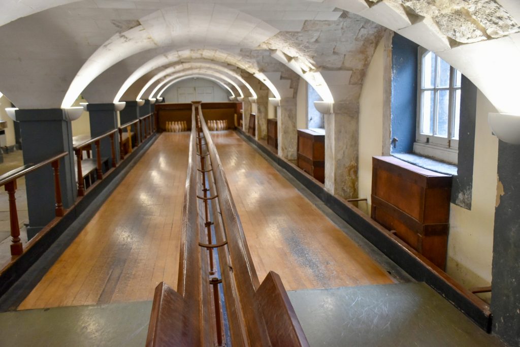 Looking down a wooden bowling alley lane with rows of wooden skittles at the far end. White Vaulted stone roof over the top
