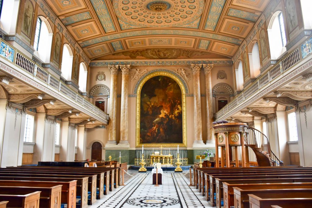 Looking towards the alter of a large square space with brown wooden pews at the bottom on either side of a white and black tiled floor. Balconies on either side and beautiful painted pink and green photo. Gold alter pieces and an old painting above the alter