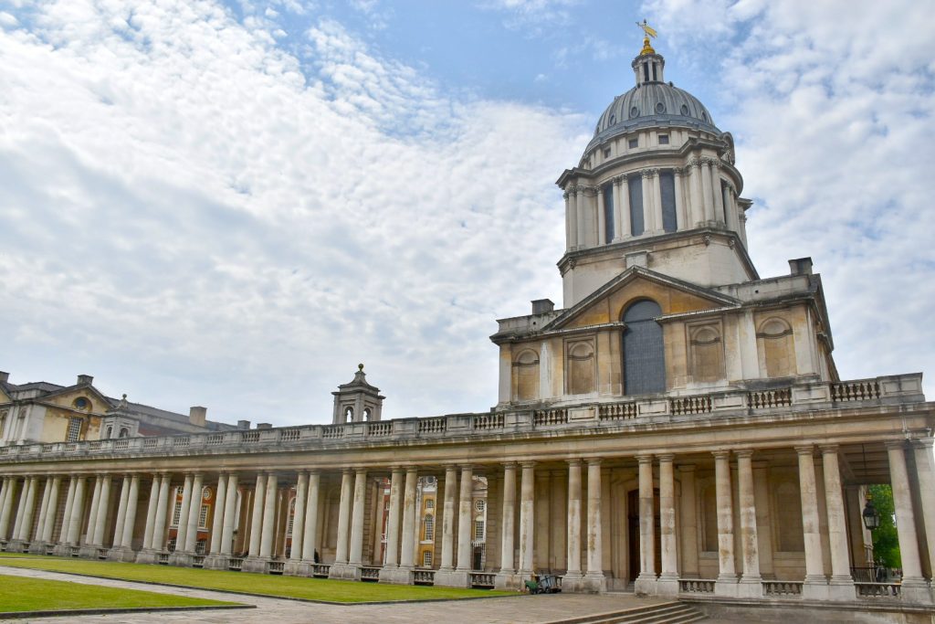 Photo of an impressive stone building running across the bottom - it has rows of columns along the length of the building, and a domed tower above. Cloudy sky behind