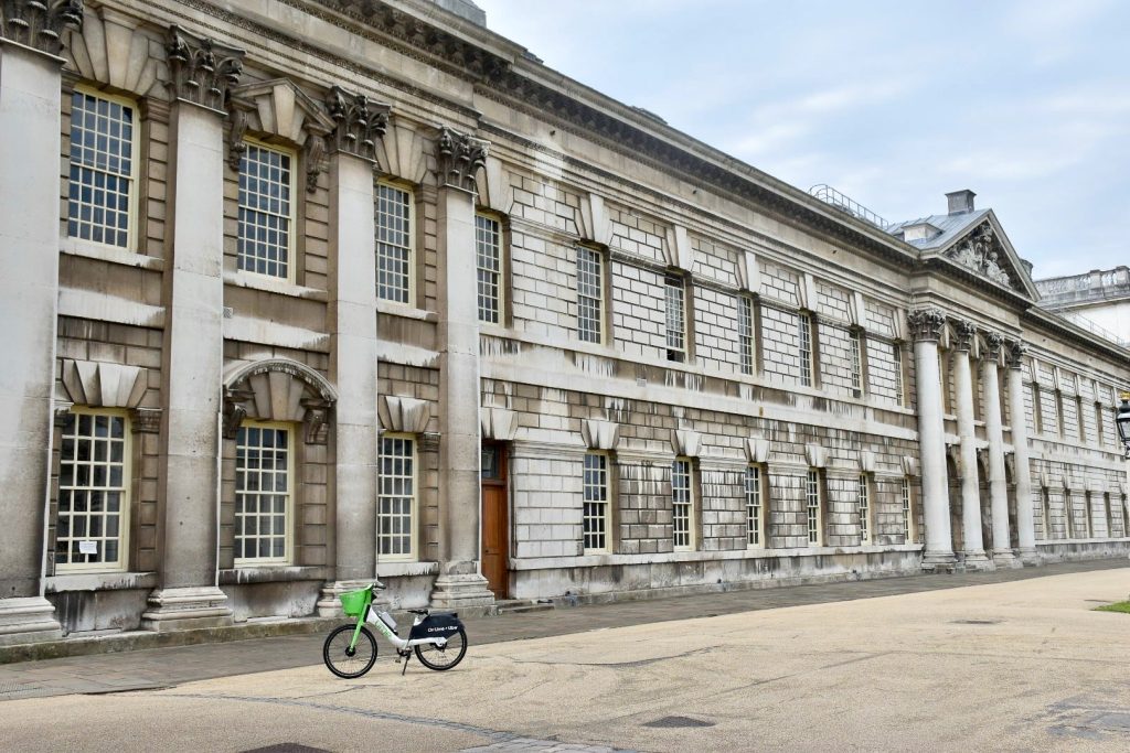 long white stone building with columns which reaches from one side of the photo to the other. It has rows of square pane windows over two flours. Grey sky above. Green and White City bike to the bottom left
