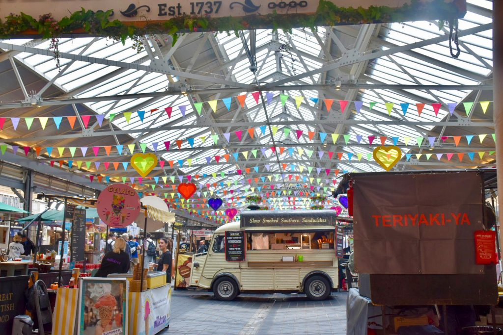 Looking into a large structure with glass panelled ceiling with rows of bunting along the length of the ceiling. At ground level, there are food vans and stands - Gelato stand to the left, Teriyaki stand to the right and Slow Smoked Southern Sandwiches van straight ahead