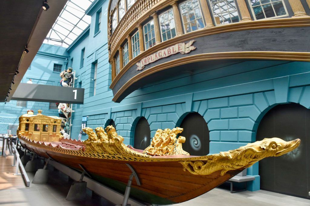 Turquoise painted brick wall in the background with curved windows that look like an old fashioned boat overlooking the floor of a museum. In the foreground is a long wooden barge with ornate gold trimmings (lions and fish) and a covered cabin at the far end