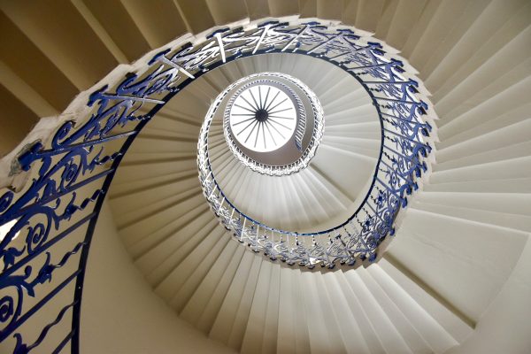 Looking upwards through the centre of a white stone spiral staircase. The banister is wrought iron, painted royal blue and ornate, featuring a tulip design