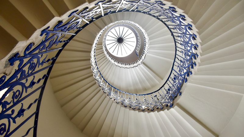 Looking upwards through the centre of a white stone spiral staircase. The banister is wrought iron, painted royal blue and ornate, featuring a tulip design