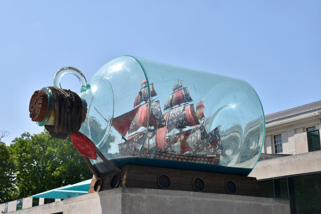 Looking upwards at a large glass bottle enclosing an old sailing ship (ship in a bottle) facing to the left. It sits on a stone plinth in front of a building with blue sky behind
