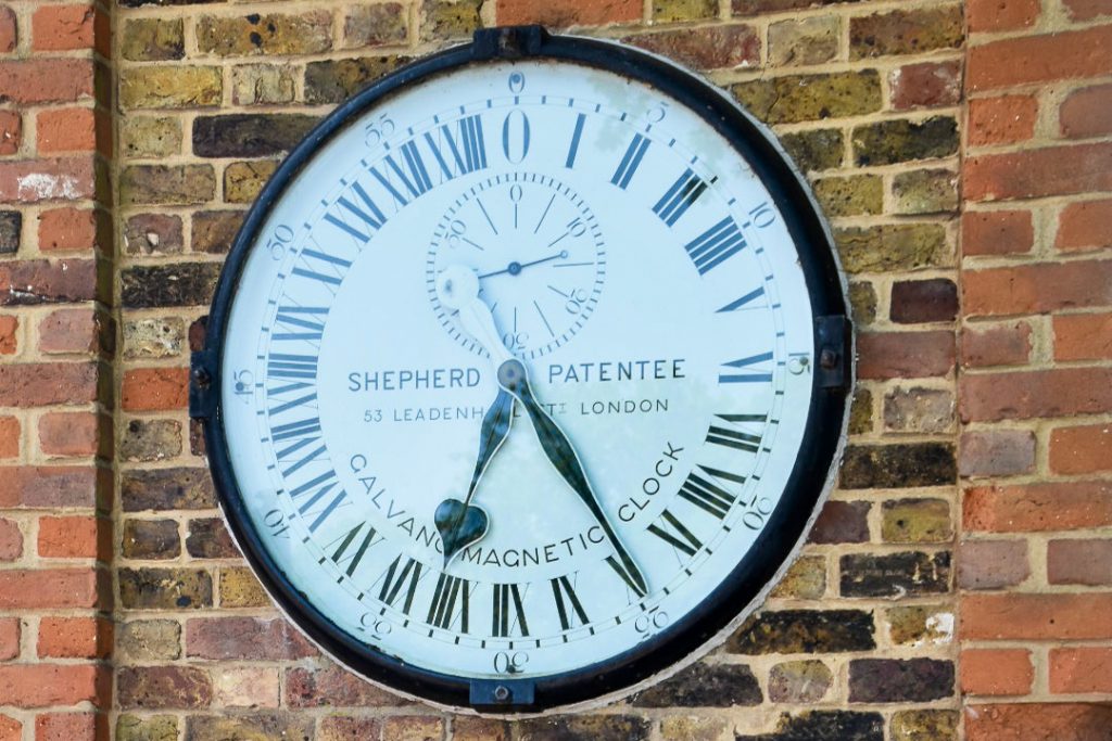 A large circular clock face is mounted in a brick wall. It has a black trim and hands on a white background. It says Shepherd Patentee of Leadenhall, London and Galvano Magnetic Clock