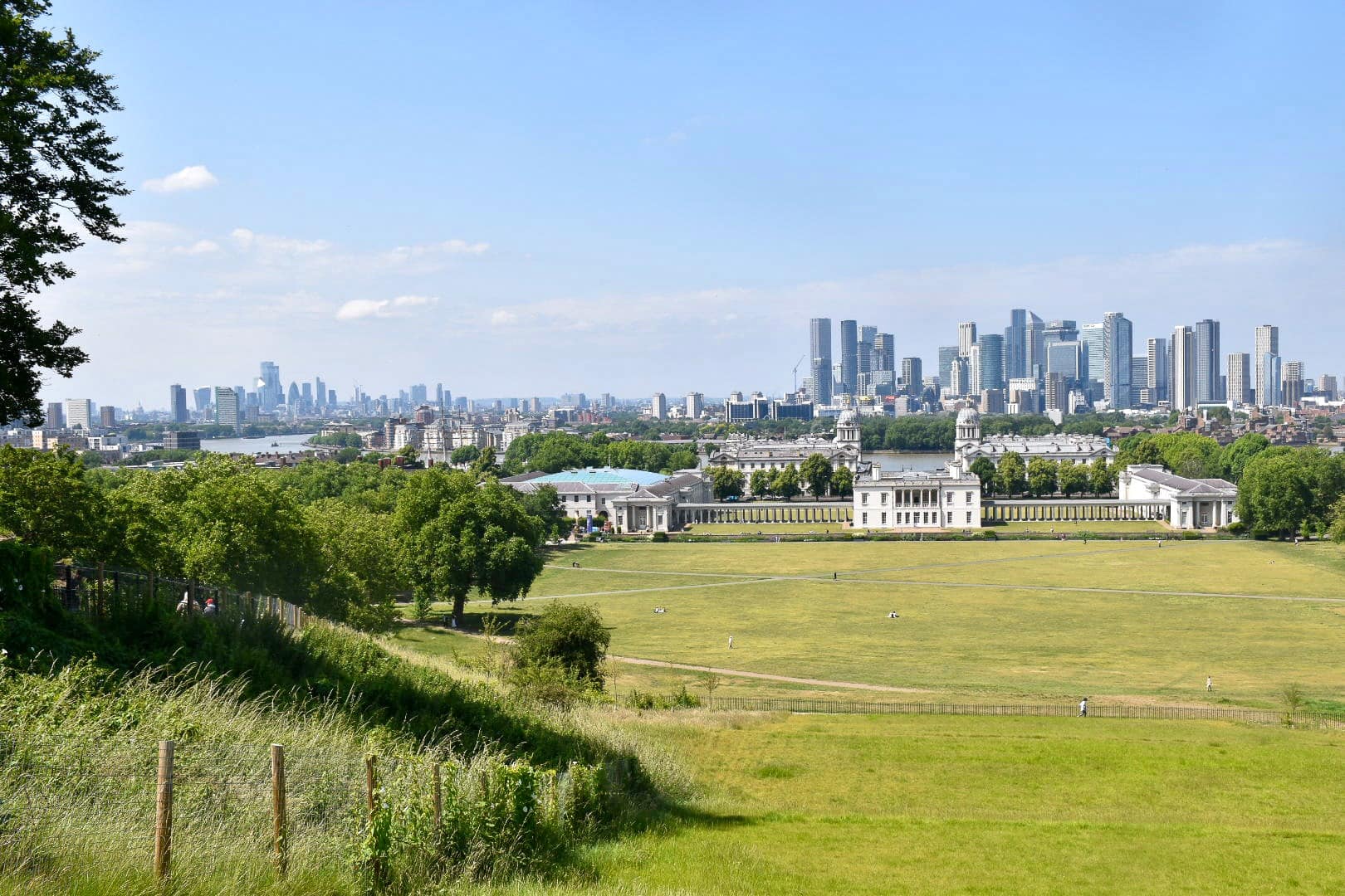 View from the top of a hill looking down a sweeping green lawn, trees to the left, to a complex of white stone buildings on the horizon, with tall office blocks behind. To the left of the photo you can just see a river winding through the office blocks