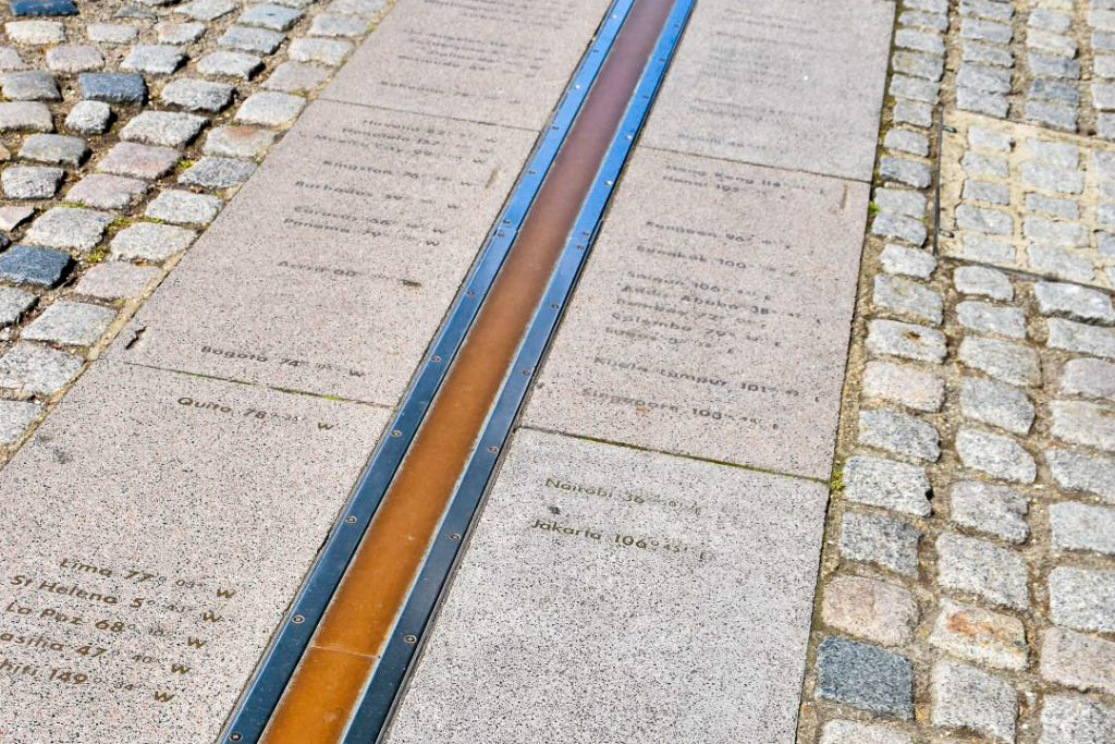 View of the pavement with small square bricks to the left and right of a paved section in the middle. Running along the centre is a bras strip trimmed by metal. There are the names of cities with their degrees of latitude printed in the pavement
