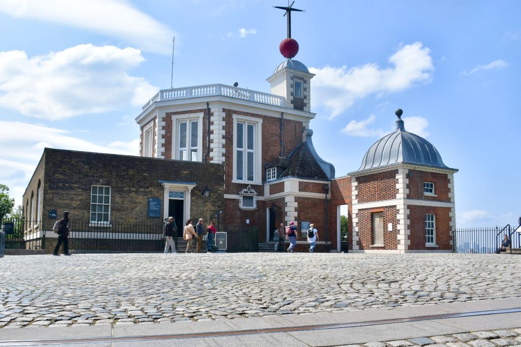 View of an old fashioned red brick building in the top half of the photo. The building has white trim on the sides and there is a red ball and weathervane on the top of the highest tower. Pale blue sky behind. Cobbled area in front of the building a paved strip running along the bottom of the photo
