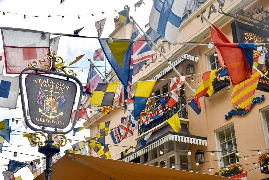 Photo of the rows of flags outside the Trafalgar Travern pub in Greenwich - there is a pub sign to the left on freestanding back sign, and peach coloured pub building just visible to the right behind the chaotic arrangement of flags. The flags are mainly shipping flags, with a couple of Union Jacks