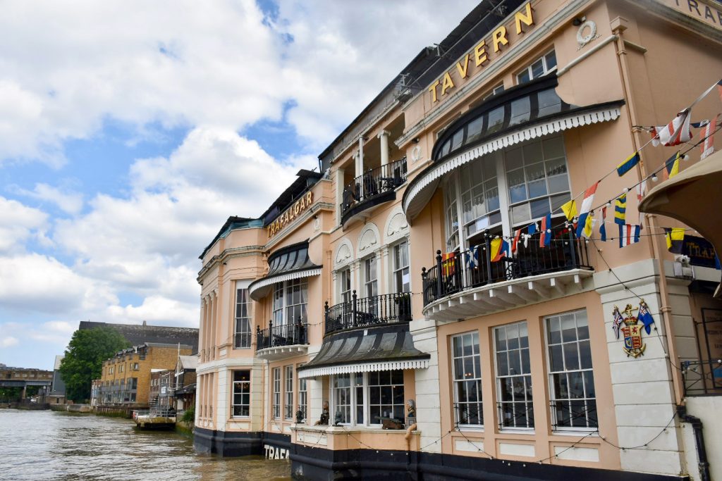 View of a salmon pink painted pub biuilding with back verandas and white window panes, overlooking a river. It has the wording Trafalgar Tavern in gold letters at the top and rows of flags underneath it