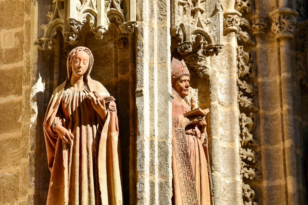 Two stone figures are carved into the corner of what appears to be a church building. The left is a lady and the right a male priest in robes. Both appear to be ready from books.