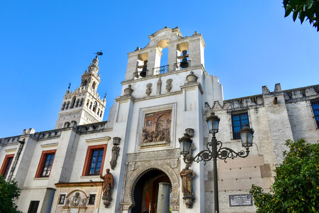 Looking up at a white stone facade covered in stone statues around an arched doorway. There are bells on top of the entrance way, an ornate black lamp post in the foreground and blue sky above. Behind the building is a tower in Moorish design