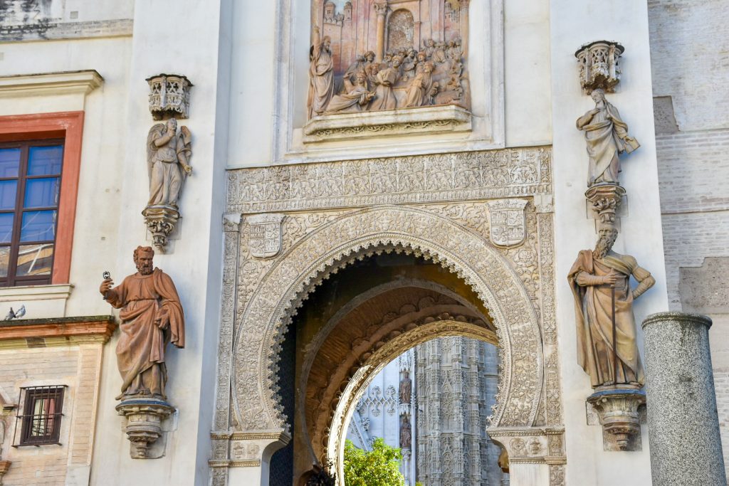 A white wall fills the frame with a horse-shore shaped arch in the centre decorated in Islamic designs. Either side of the arch are stone statues mounted onto the walls. Above the arch is a stone frieze featuring figures in front of a building.