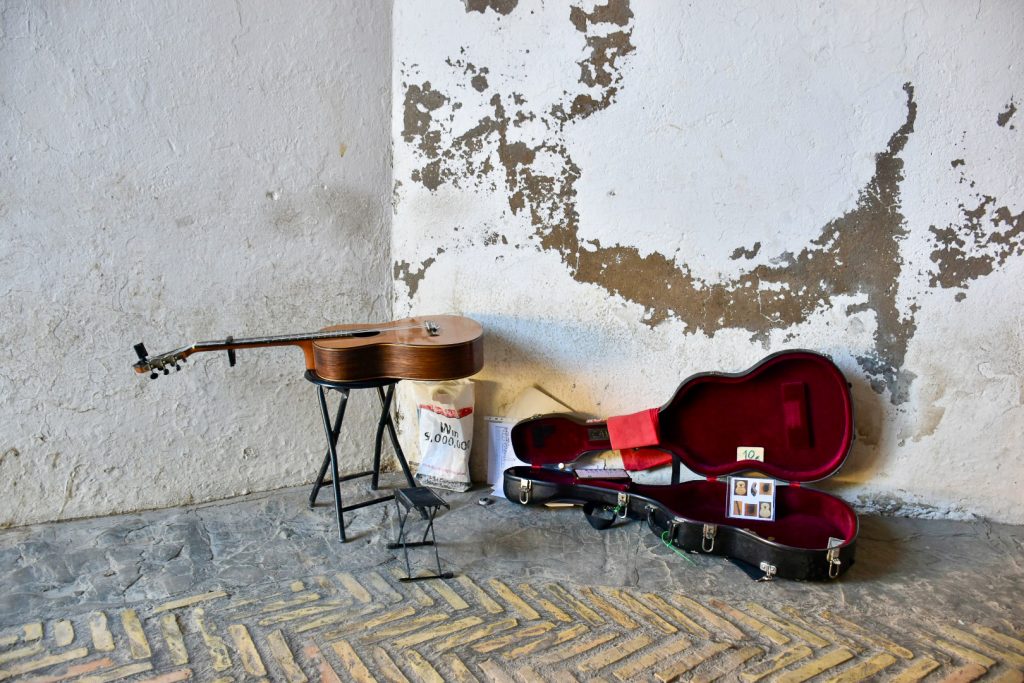 An acoustic guitar sits on a metal stool in front of an old painted corner wall. To the right hand side, an open guitar box sits on the floor with a red velvet interior. Floor tiles are laid at diagonals.