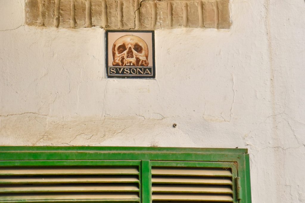 White plaster wall. Bottom shows a green slatted window. Above is a plaque featuring the painting of a skull and the word Susona.