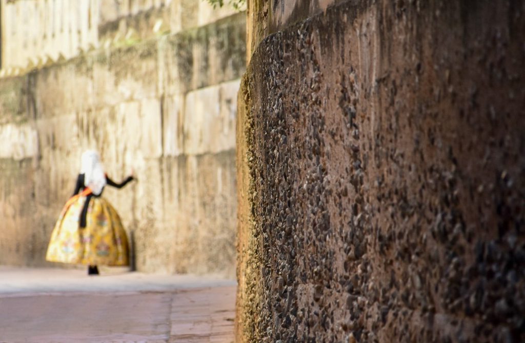Right hand side of the photo is an old brown wall, Left hand side of the photo looks down the length of the wall where the fuzzy outline of a lady in old fashioned clothes (white headdress, black top and gold hooped skirt) leans on the wall