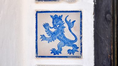 White stone wall with blue and white tiles inserted. The tile in the centre of the photo features a lion in a crown. Dark wood doorway to the right hand side