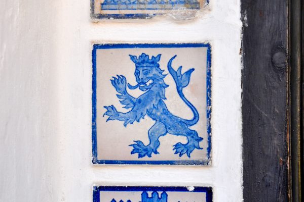 White stone wall with blue and white tiles inserted. The tile in the centre of the photo features a lion in a crown. Dark wood doorway to the right hand side