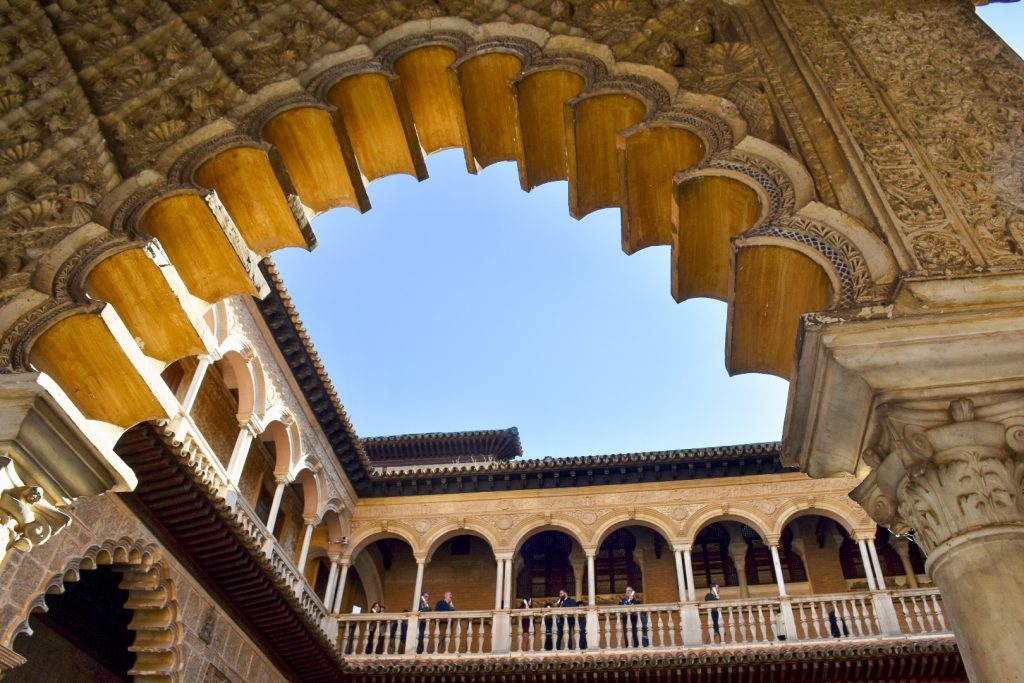Looking upwards through a Moorish arch to the pale blue sky beyond. A row of arches is visible around what appears to be a square