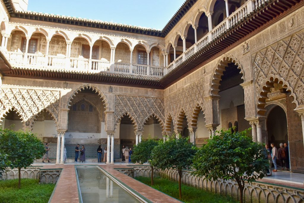 Inside a courtyard with Moorish styled arches around a channel of water lined by shrubs. The buildings have a second tier above with Renaissance style arches.