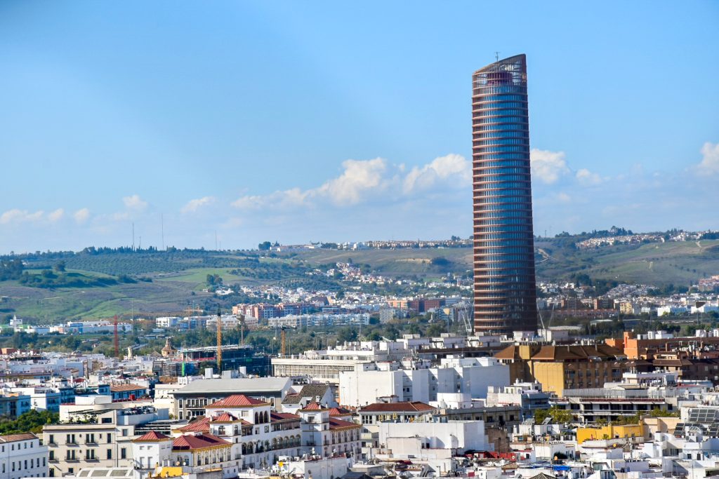 City scape taken from up high with hills in the background and pale blue sky above. To the right of the centre is a tall circular modern skyscraper (shaped like a lipstick)