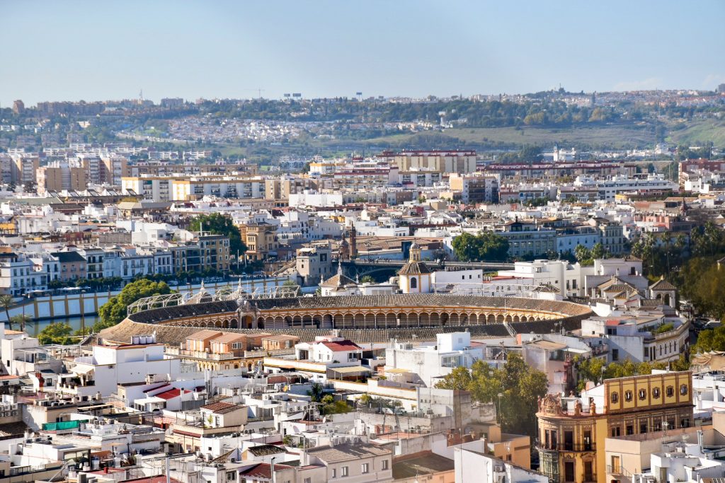 Scene from on high looking down on a city fulll of white low rise buildings. Hills in the background and pale grey sky above. In the centre of the photo you can see the large oval shape of a bull ring
