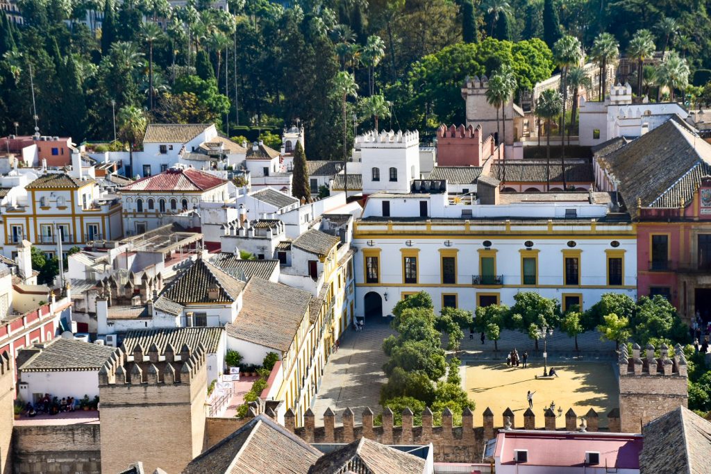 View from above looking down on buildings in a town. The buildings are low rise and mainly white or beige. A courtyard can be seen to the bottom right with yellow window frames on white walls and trees around the square. trees in the background