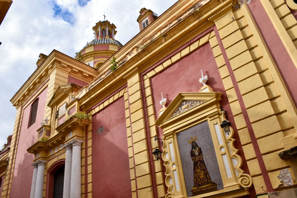 Red and yellow painted church with religious painting on the outside. White clouds and blue sky
