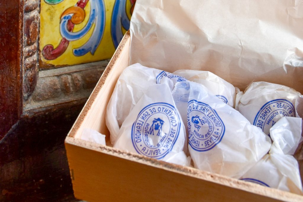 An open wooden box sits to the bottom right hand side of the frame with signs of painted tiles to the left. Inside the box are a number of circular items wrapped in white greaseproof paper with blue lettering printed on.