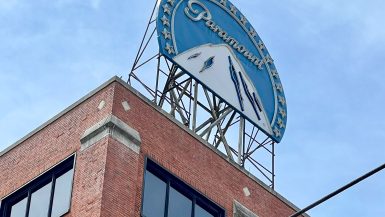 Old fashioned blue curved sign with a white mountain and the word Paramount sits on top of a red brick building. Pale blue sky behind.