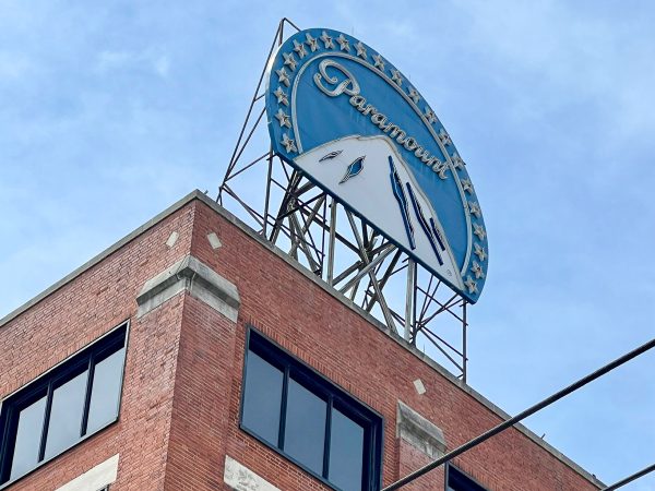 Old fashioned blue curved sign with a white mountain and the word Paramount sits on top of a red brick building. Pale blue sky behind.