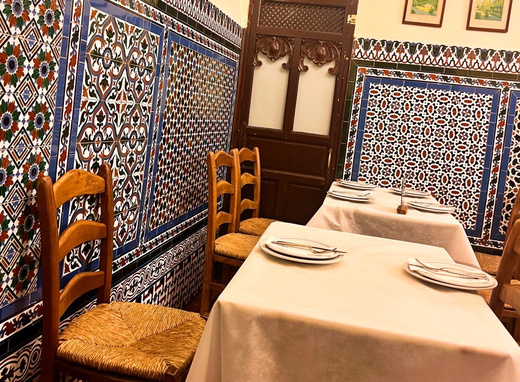 Inside a restaurant with tiles on the walls featuring geometric designs in blue which and red colours. There is a wooden doorway in the middle of the frame. There are two tables with white table cloths and brown whicker chairs, ready for diners