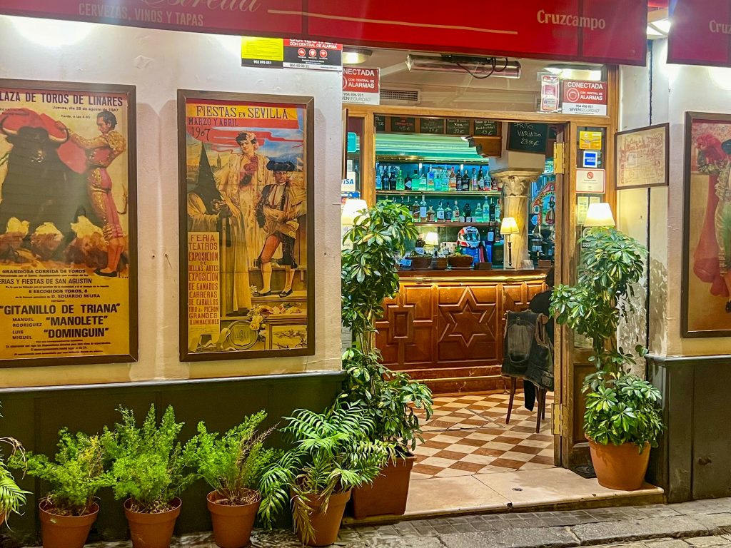 The front of a bar or cafe with green potted plants in front and framed prints relating to bull fighting on the walls. You can see into the building which has a wooden bar behind!