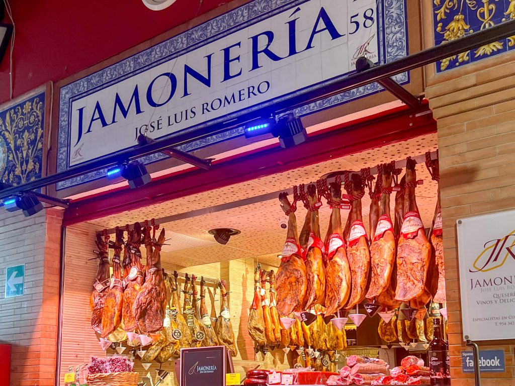 Market stall with hanging legs of ham on display and a tiled header over the shop window saying Jamoneria (Jose Luis Romero)
