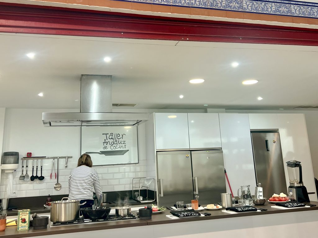 Looking into a kitchen with white units and silver appliances. In the middle is a work station for a kitchen. The name says 'Taller Andaluz de Cocina'