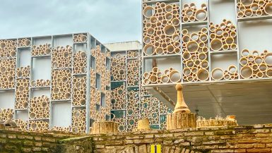 A modern building with metal frames filled with circular bubble designs in yellow stone over a stone bridge archway.