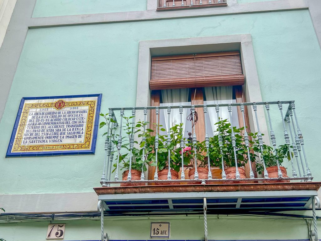 A minty green wall fills the frame with a shuttered window behind a wrought iron balcony with plant pots on it. Tiled plaque to the left hand side