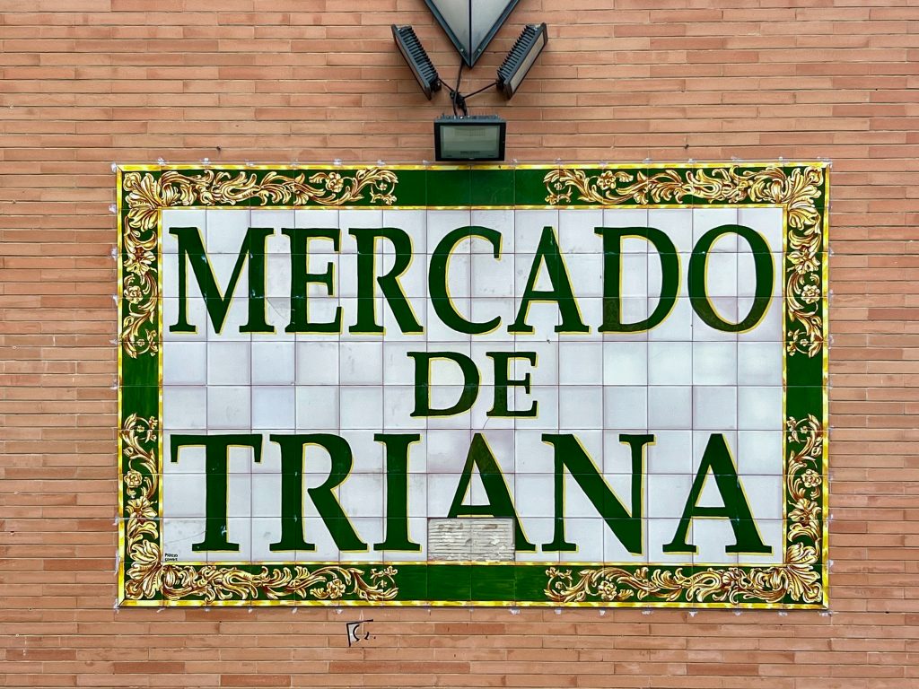 Brick wall with a sign saying Mercado de Triana made out of tiles. Dark green letters on white tiled backgrounds. Border is dark green with yellow ornate decoration