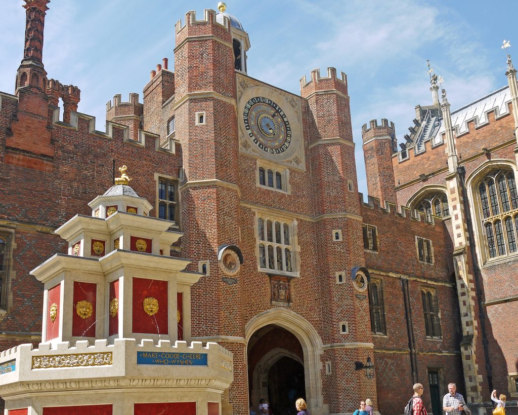 Red brick palace in Tudor style with a white and red water fountain to the left. The entrance of the palace has an ornate sundial at the top over the arched doorway