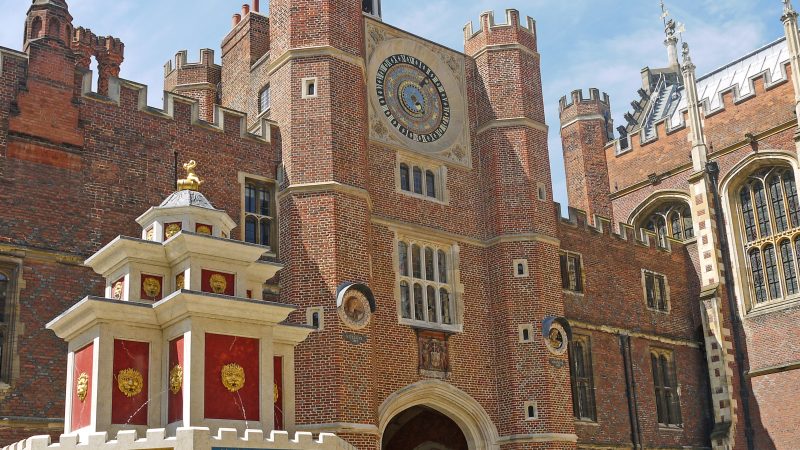 Red brick palace in Tudor style with a white and red water fountain to the left. The entrance of the palace has an ornate sundial at the top over the arched doorway