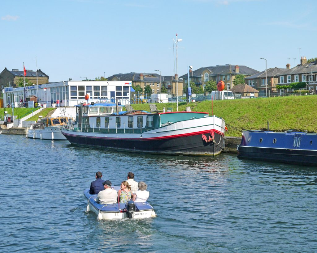 River seen with a small motor boat in foreground with 5 passengers. A large barge (green, white and black) plus other boats are moored by the river bank. Residential houses in the background