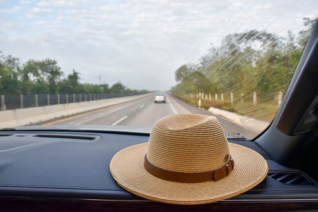 A straw hat with brown leather band sits on the dashboard of a car travelling along the road. Car in front and trees to the right