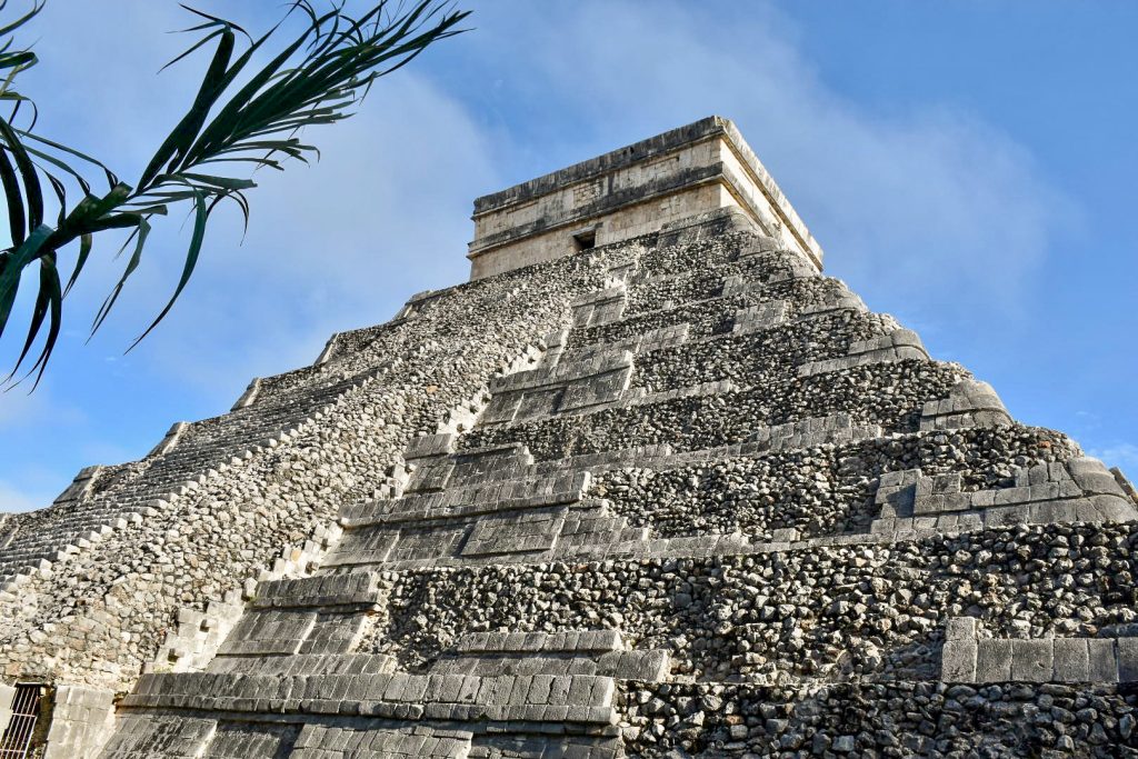 A stone pyramid temple fills the frame with blue sky behind and green leaves to the left