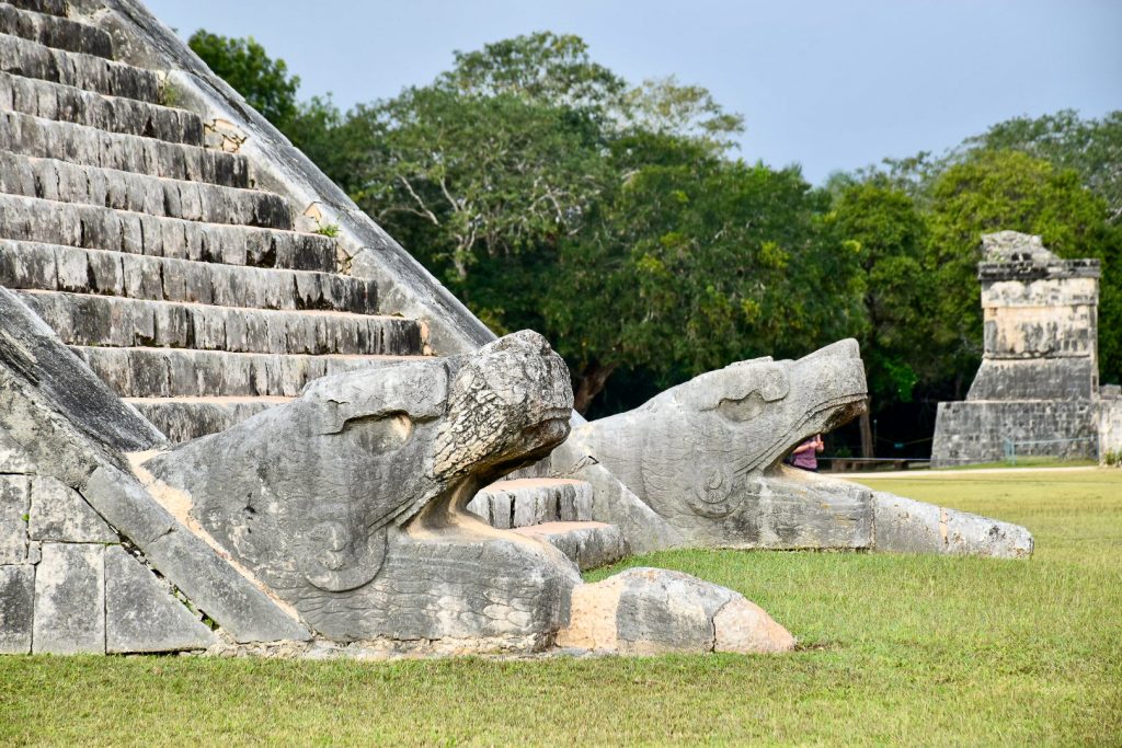 Carved serpent heads at the bottom of a stone staircase. Trees and another old building behind. Grass in front