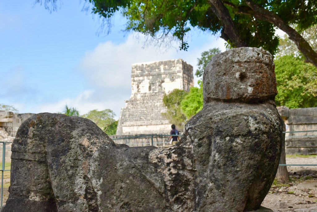 A stone figure reclines on their back - it is a worn statue and no face is evident. A stone temple like structure in the distance with trees to the right