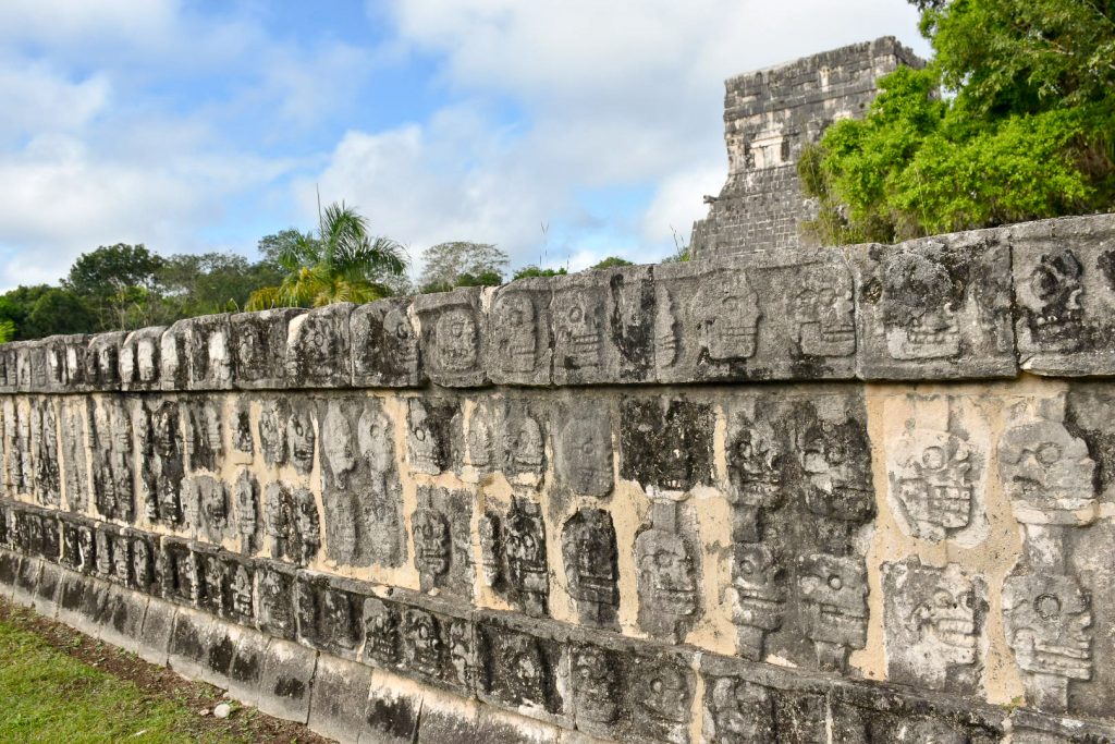 A long stone platform featuring rows of carvings of skulls. Trees and a temple behind